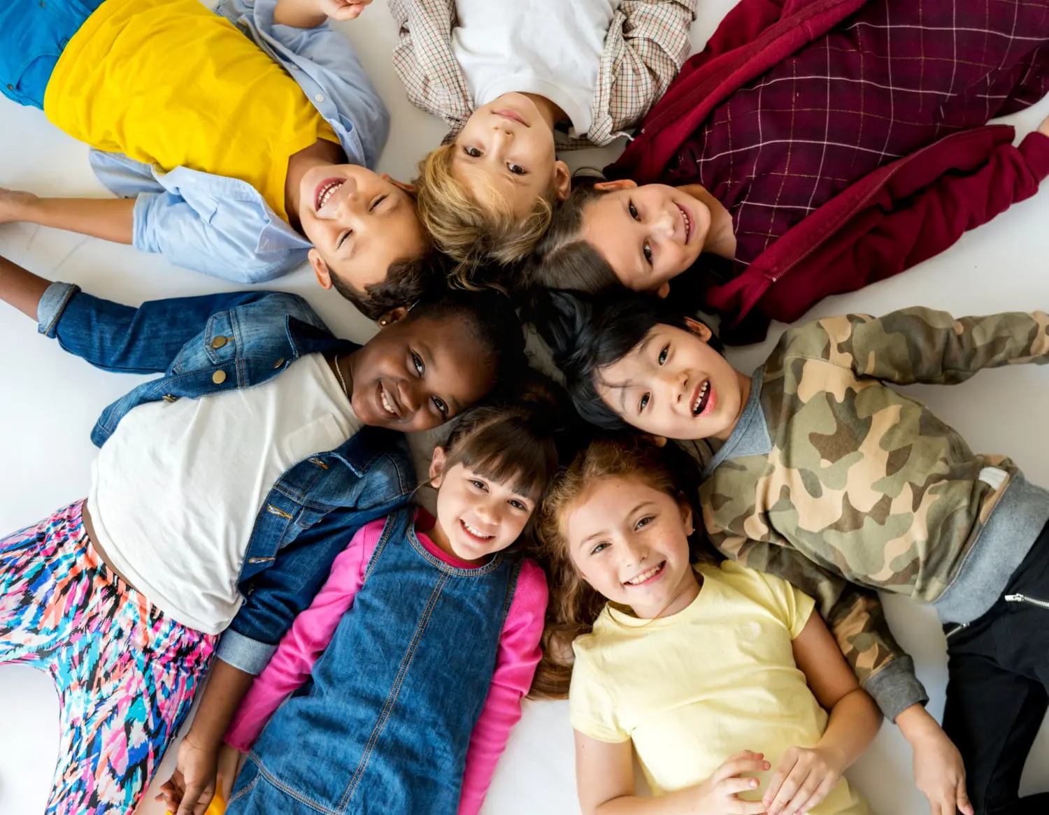 Capturing the joy of confident smiles, six children lie on floor in a circle, heads together and laughing—a perfect example of the results families can expect from age-specific pediatric dental care at Pediatric Dentistry and Orthodontics of Forsyth - Cumming in Cumming, GA.