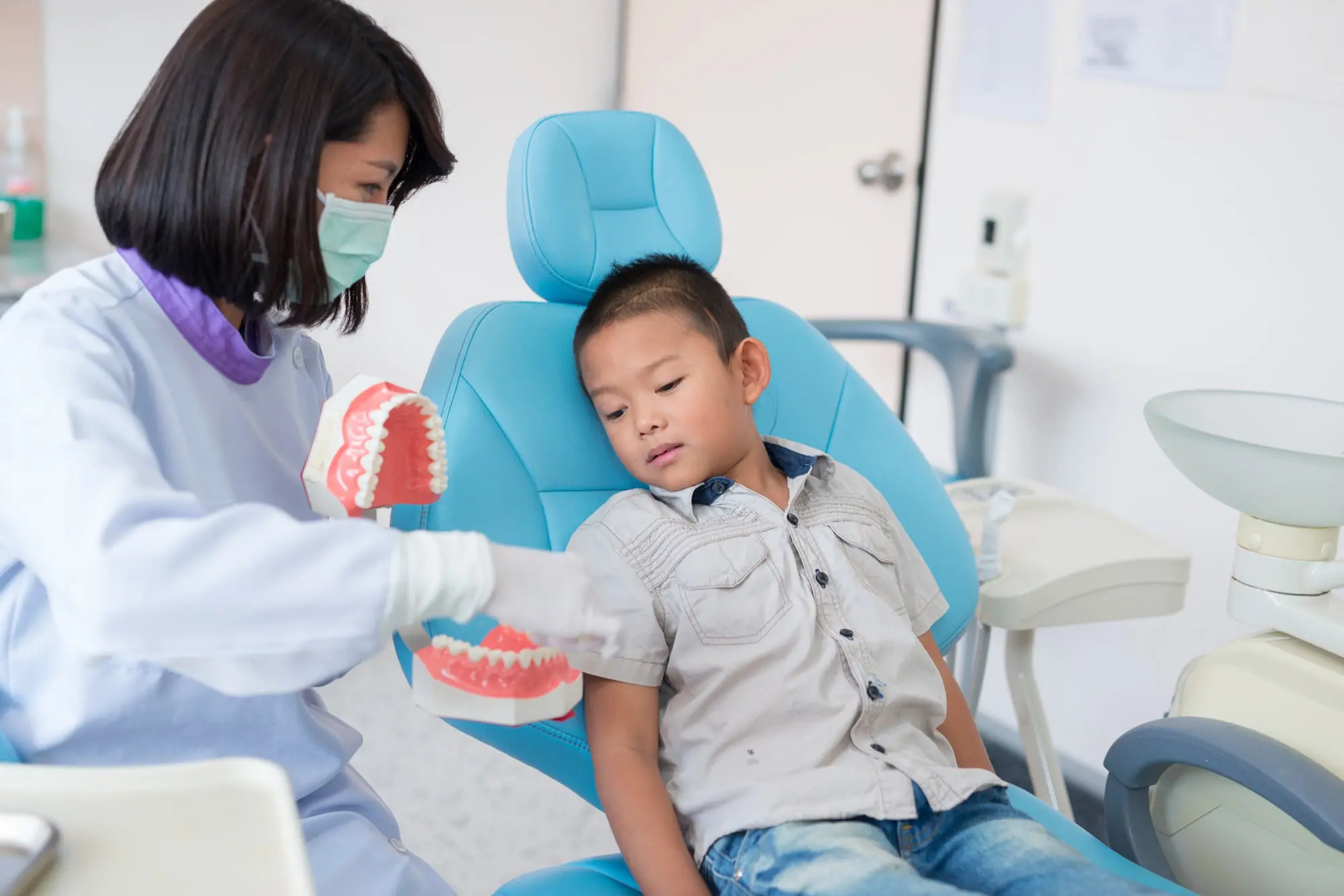 A Friendly Dentist in a white coat uses Social Stories and visual supports to explain dental care with a model to a smiling young boy in a welcoming office in Cumming, GA.