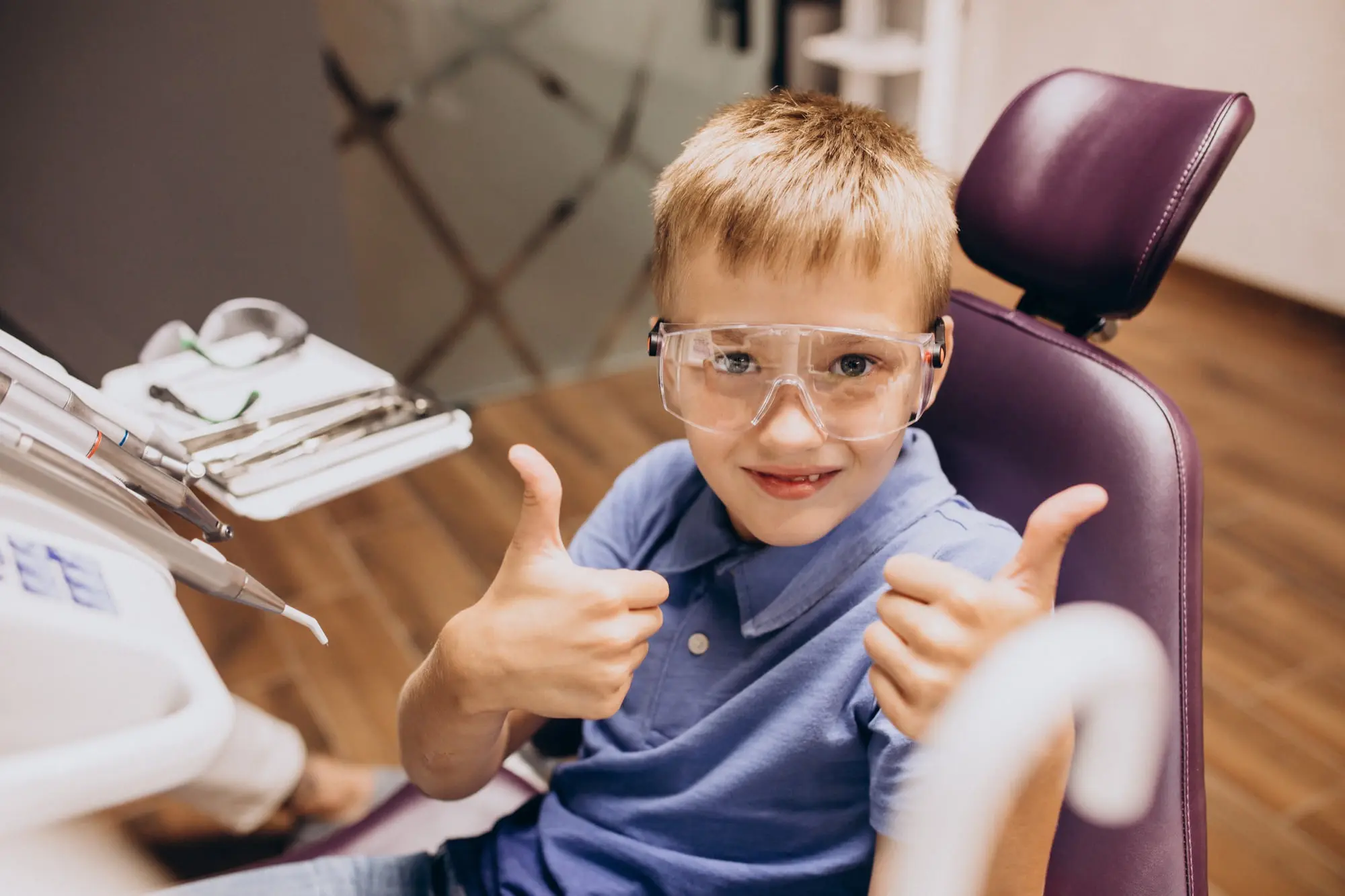 At Pediatric Dentistry and Orthodontics of Forsyth - Cumming in Cumming, GA. A young boy wearing safety glasses sits in the dental chair and gives two thumbs up, illustrating how reduced anxiety leads to more smiles at every visit for Laser Dentistry.