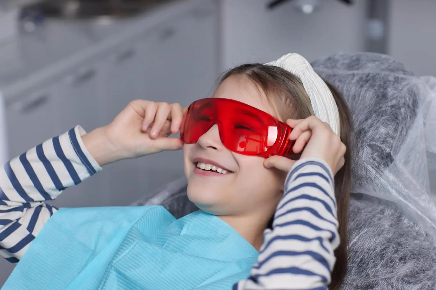 At Pediatric Dentistry and Orthodontics of Forsyth - Cumming in Cumming GA, a young child sits in the dental chair wearing a bib and red protective glasses, smiling confidently and ready to go back to school the same day with a bright, healthy smile after Laser Dentistry.