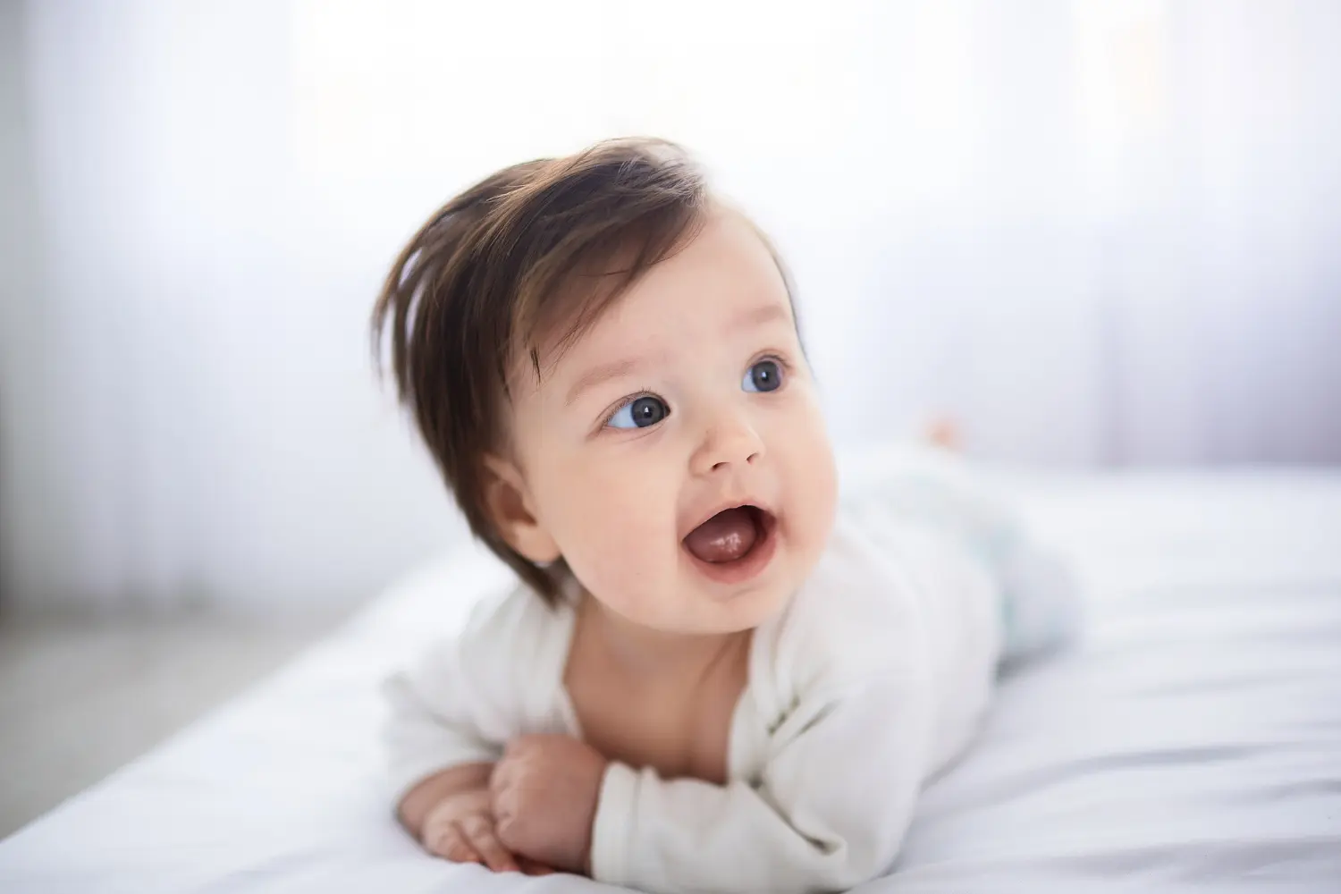 A baby smiles while lying on a bed, bathed in natural light—a heartwarming moment to cherish before their first visit to Baby Dentist in Cumming, GA by age 1.