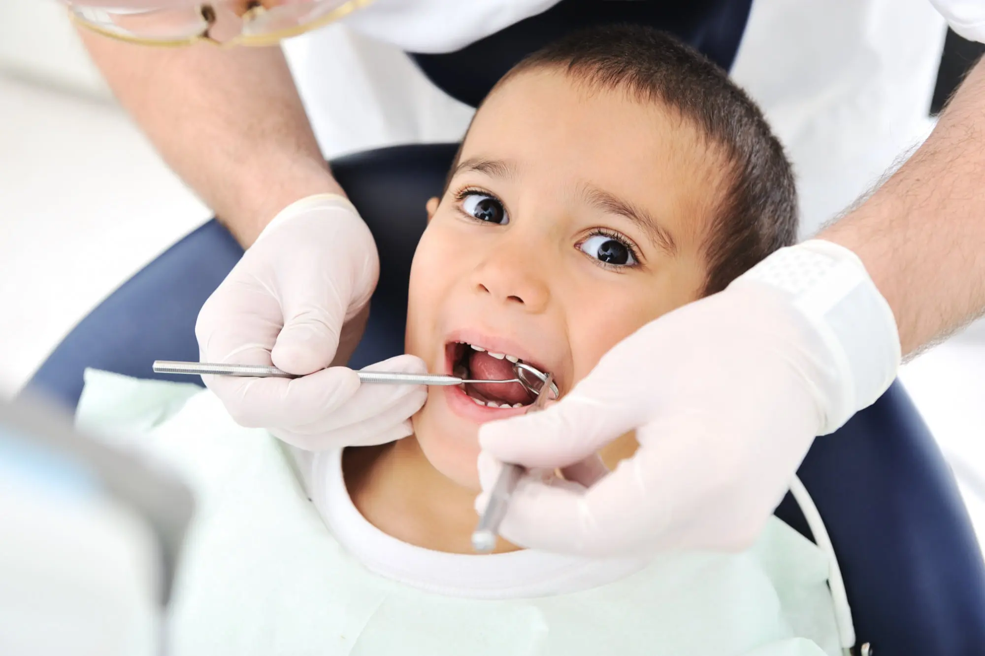 At Pediatric Dentistry and Orthodontics of Forsyth - Cumming in Cumming GA, a dentists specializing in emergency pediatric care gently examine a young boy teeth as she sits in the dental chair wearing a bib, gloves, and safety glasses are used to ensure a safe and comfortable experience.