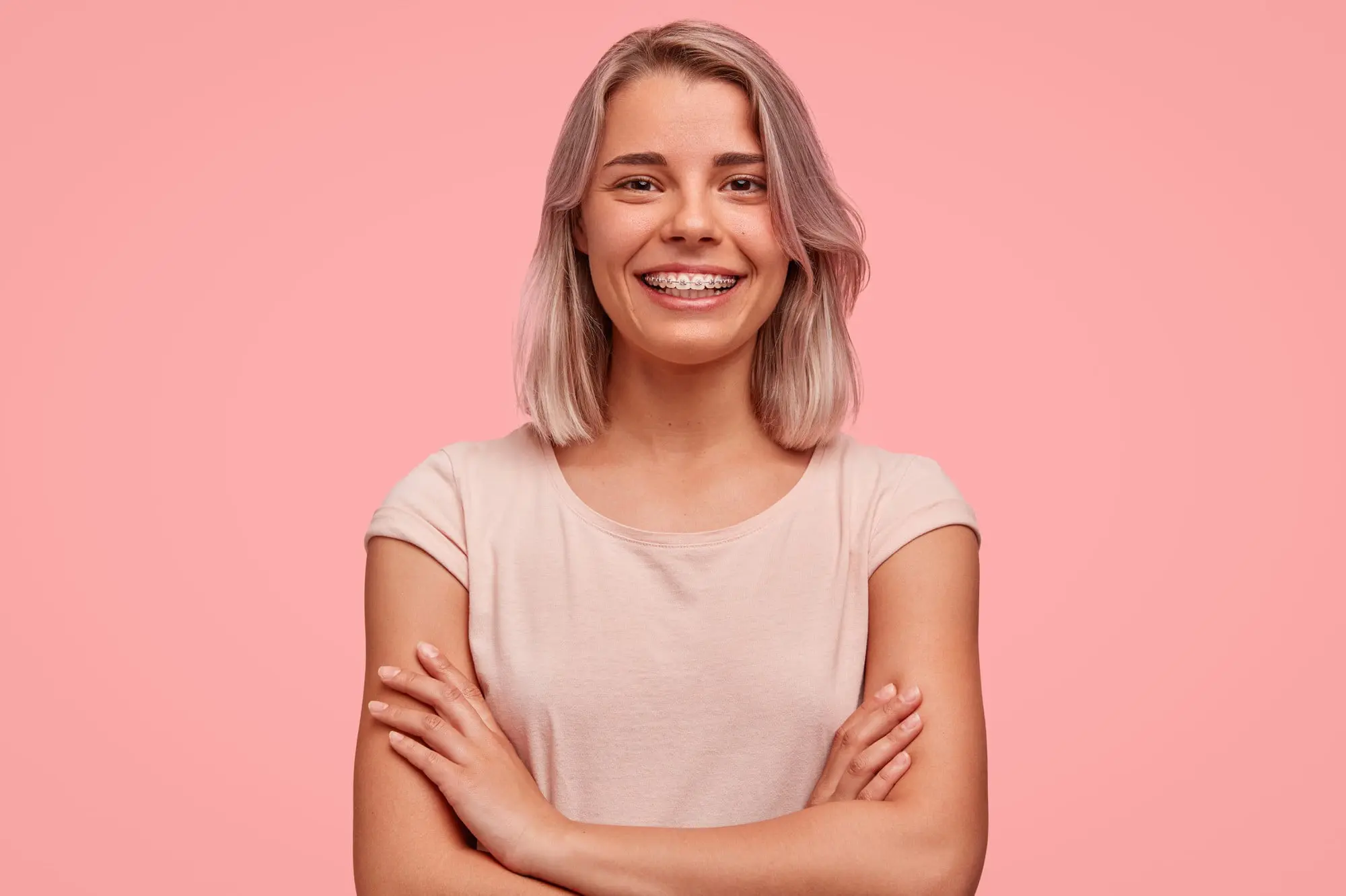 A smiling young woman with light hair and adult braces, arms crossed, stands against pink, representing Pediatric Dentistry and Orthodontics of Forsyth - Cumming in Cumming, GA.