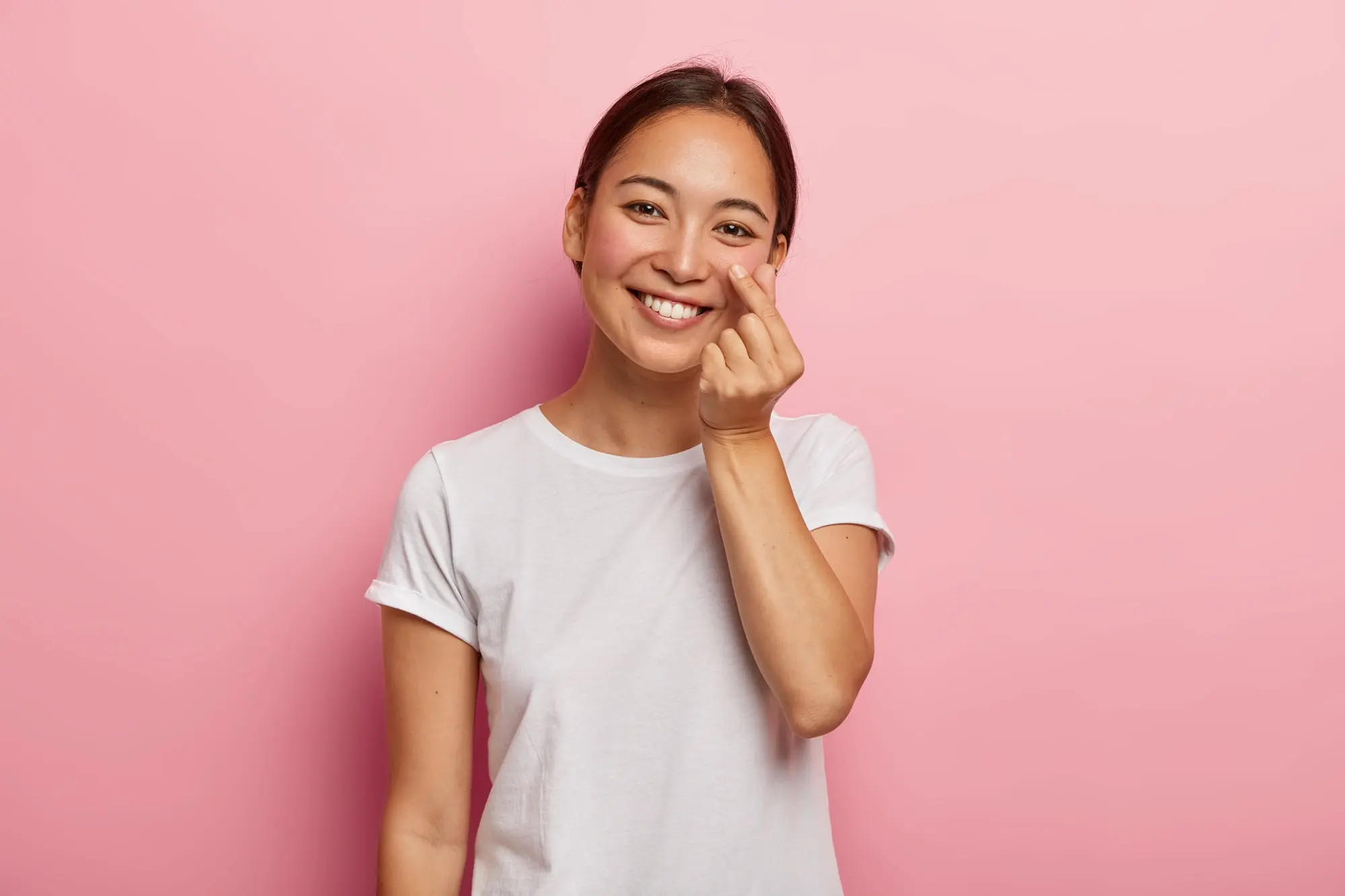 Smiling young woman in a white t-shirt makes a finger heart gesture in front of pink background represent best orthodontic appliances at Forsyth Pediatric Dentistry and Orthodontics in Cumming, GA.