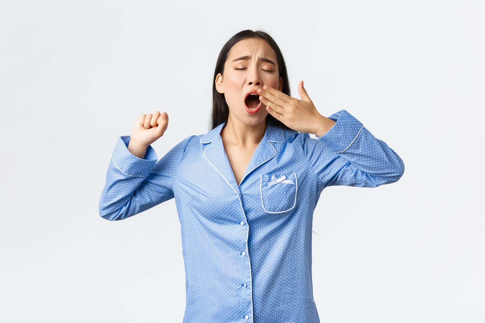 Against a plain white background, a woman in blue polka dot pajamas yawns and stretches represent mouth breathing issue in Cumming, GA.