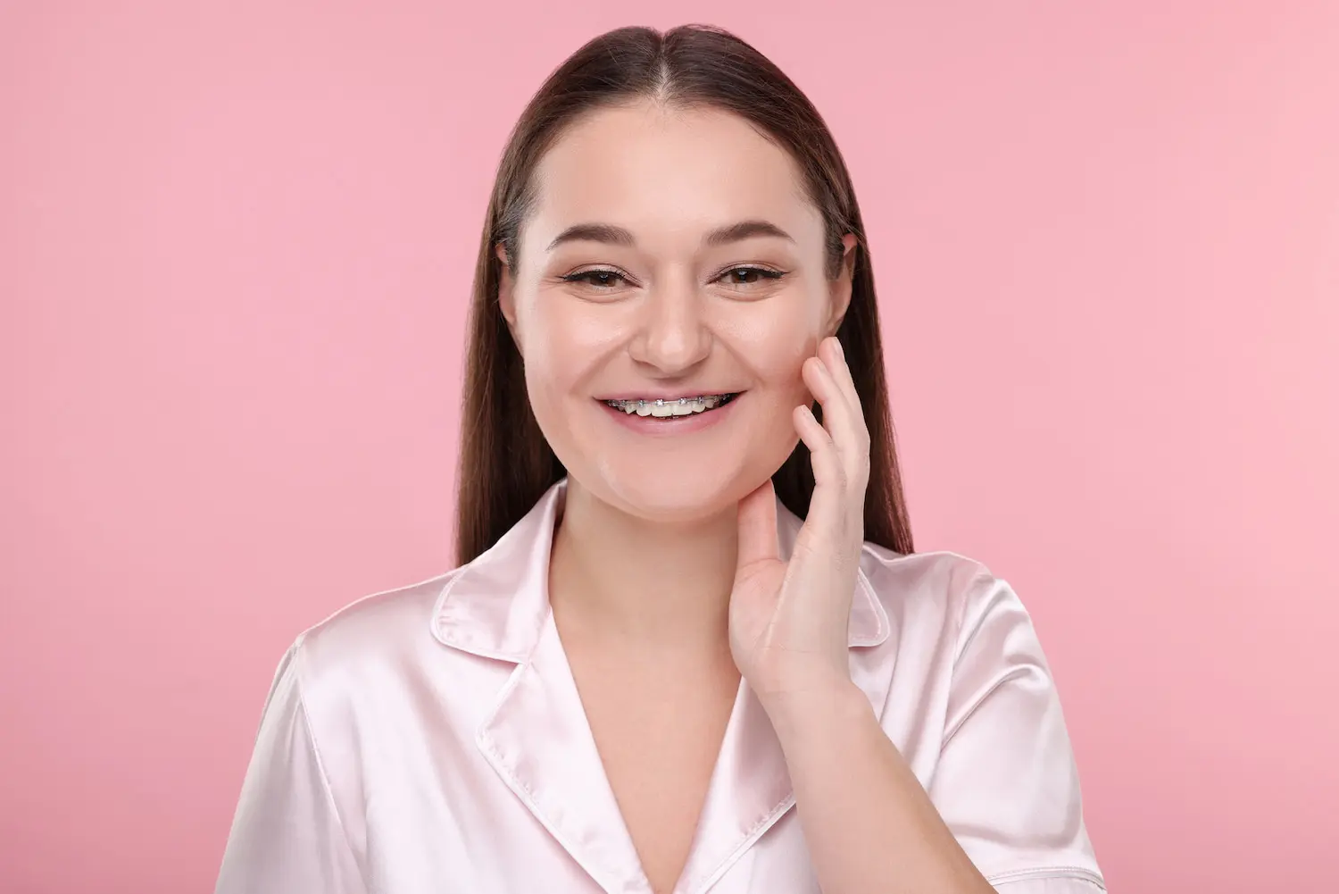 Smiling young woman with long brown hair and metal braces, in a pink satin shirt, promoting Pediatric Dentistry and Orthodontics of Forsyth - Cumming in Cumming, GA, against a solid pink background.
