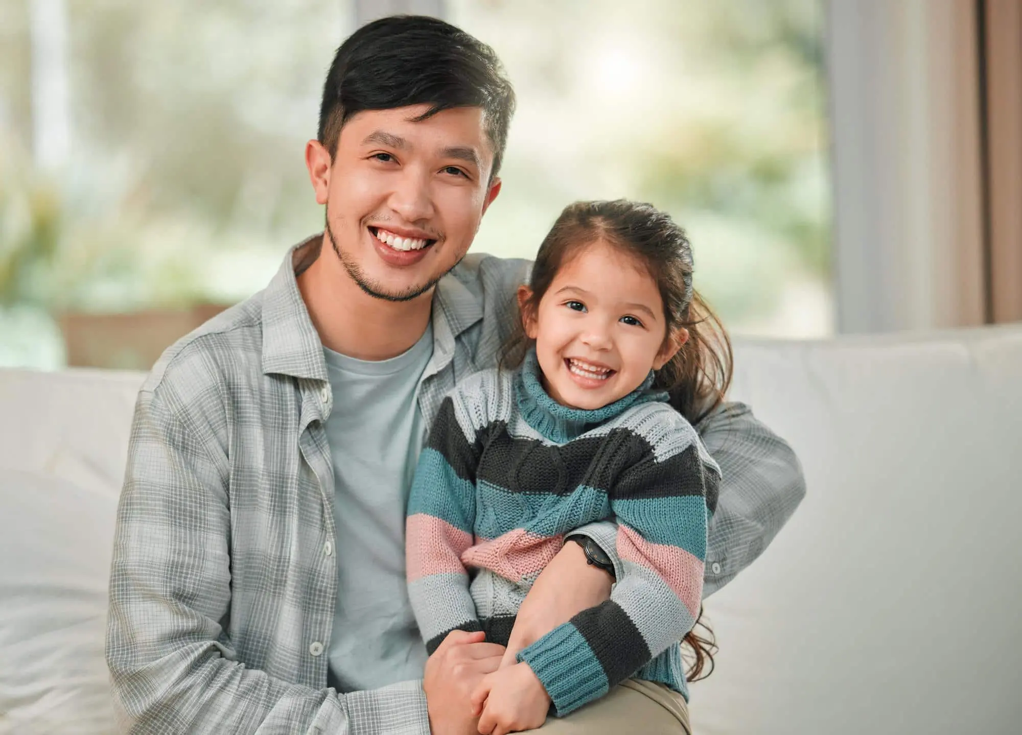 Smiling man and girl sit on a couch in a well-lit room after discussing costs at Forsyth Pediatric Dentistry and Orthodontics in Cumming, GA.