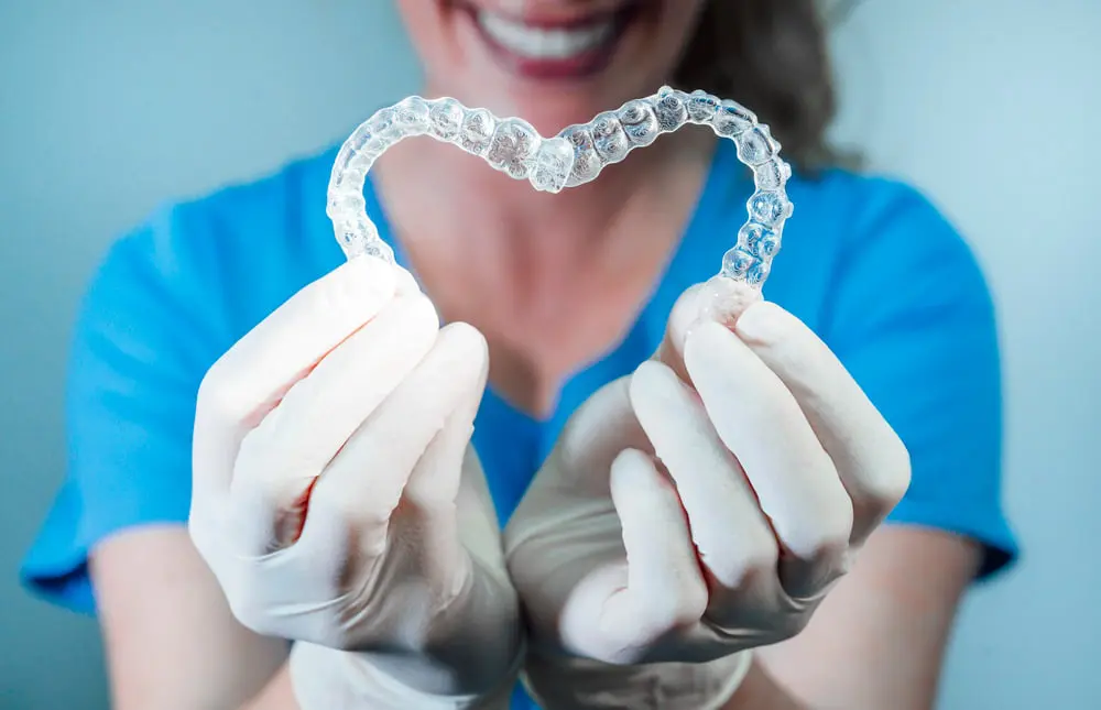 Smiling in the background, a staff member at Pediatric Dentistry and Orthodontics of Forsyth - Cumming in Cumming, GA forms a heart with Invisalign aligners.