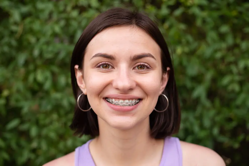 A smiling young woman with short brown hair and metal braces at Pediatric Dentistry and Orthodontics of Forsyth - Cumming in Cumming, GA. Green foliage behind.