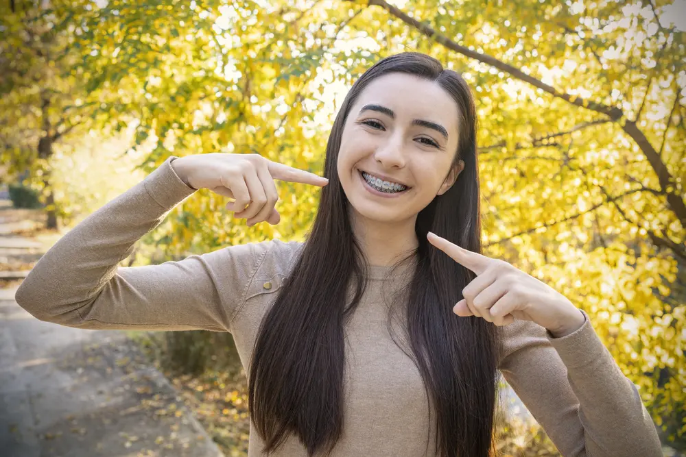 A young woman with long dark hair smiles and points to her metal braces; Pediatric Dentistry and Orthodontics of Forsyth - Cumming in Cumming, GA.