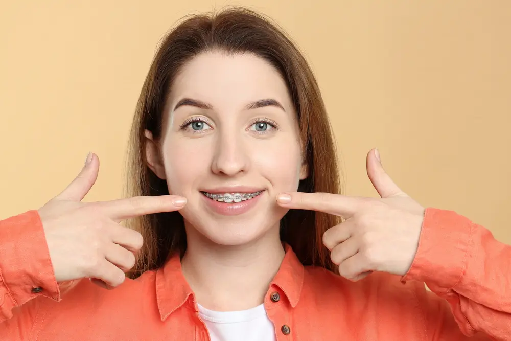 A young woman in an orange shirt points to her metal braces, smiling, representing Pediatric Dentistry and Orthodontics of Forsyth - Cumming in Cumming, GA.
