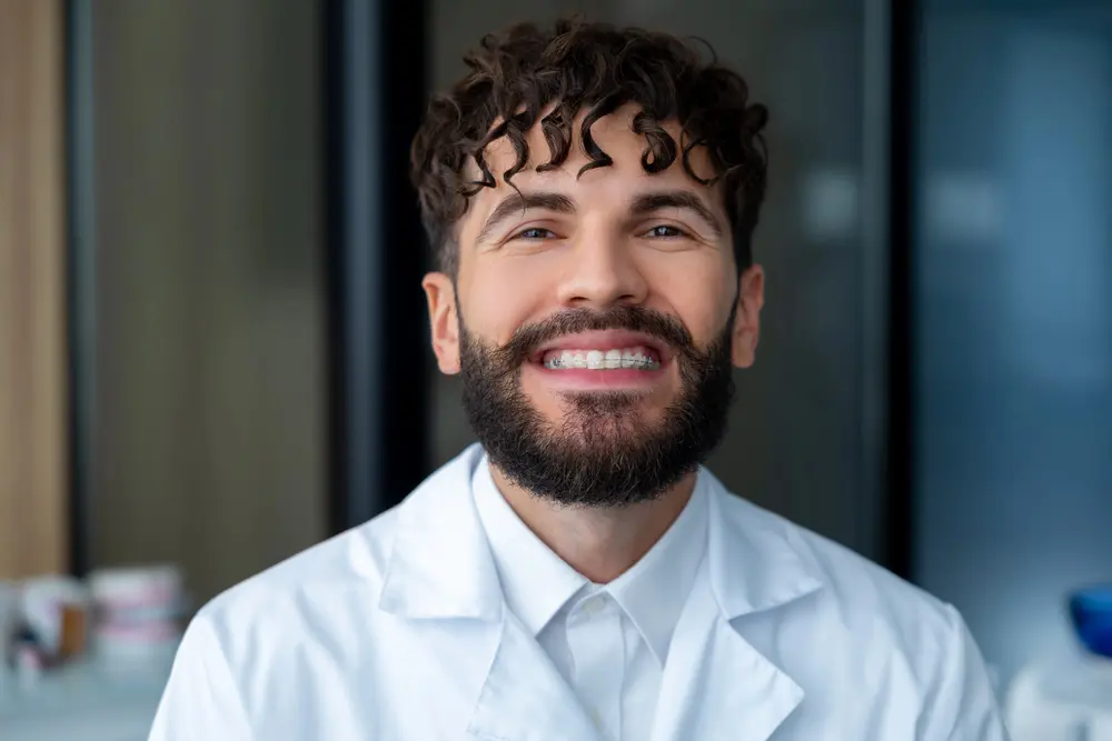 A man with curly hair and a beard in a white lab coat smiles confidently, representing Pediatric Dentistry and Orthodontics of Forsyth - Cumming in Cumming, GA with Clear Braces.