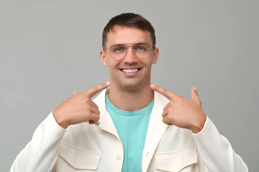 A man in glasses and a white jacket smiles, pointing to his metal braces, representing Pediatric Dentistry and Orthodontics of Forsyth - Cumming in Cumming, GA.