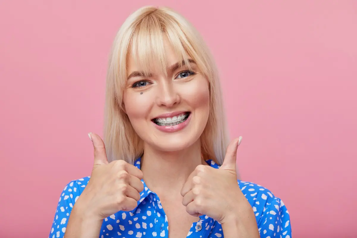 Blonde woman with clear braces in blue spotted shirt gives two thumbs up before pink backdrop for Pediatric Dentistry and Orthodontics of Forsyth - Cumming in Cumming, GA.