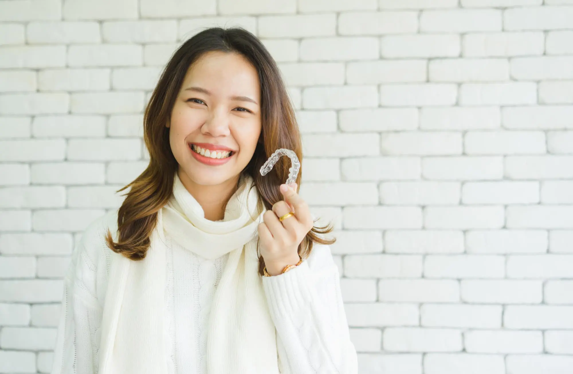 Smiling woman in a white sweater holds an Invisalign aligner at Pediatric Dentistry and Orthodontics of Forsyth - Cumming in Cumming, GA.