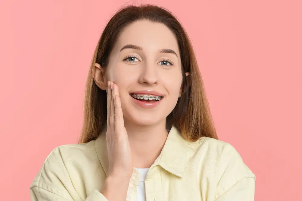 A young woman with long brown hair and metal braces smiles, touching her cheek before a pink background at Pediatric Dentistry and Orthodontics of Forsyth - Cumming in Cumming, GA.