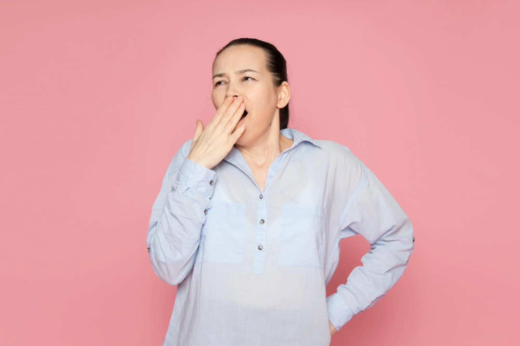 At Forsyth Pediatric Dentistry and Orthodontics in Cumming GA, a woman in a blue shirt yawns, covering her mouth against a pink backdrop.