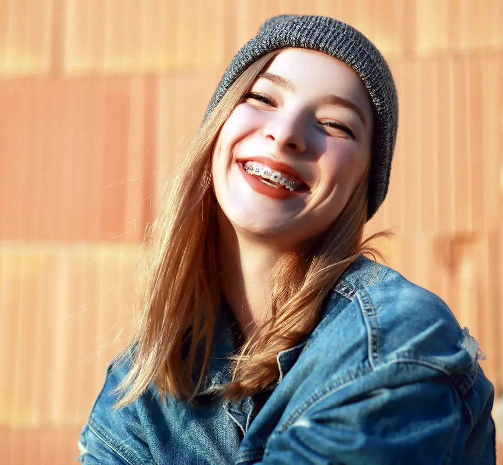 A smiling person with metal braces at Forsyth Pediatric Dentistry and Orthodontics in Cumming GA, in front of an orange wall.