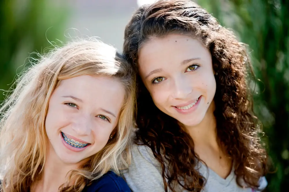 Smiling teens, one with blonde hair and braces, the other with curly brown hair, at Forsyth Pediatric Dentistry and Orthodontics in Cumming, GA.