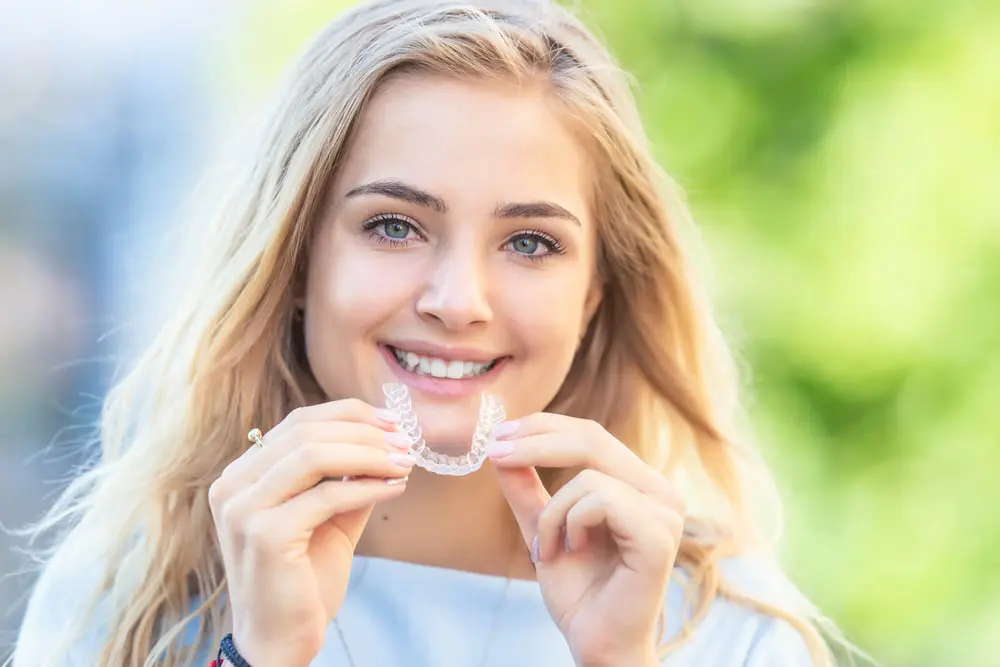 Smiling outdoors, a woman holds a clear dental aligner from Forsyth Pediatric Dentistry and Orthodontics in Cumming, GA.