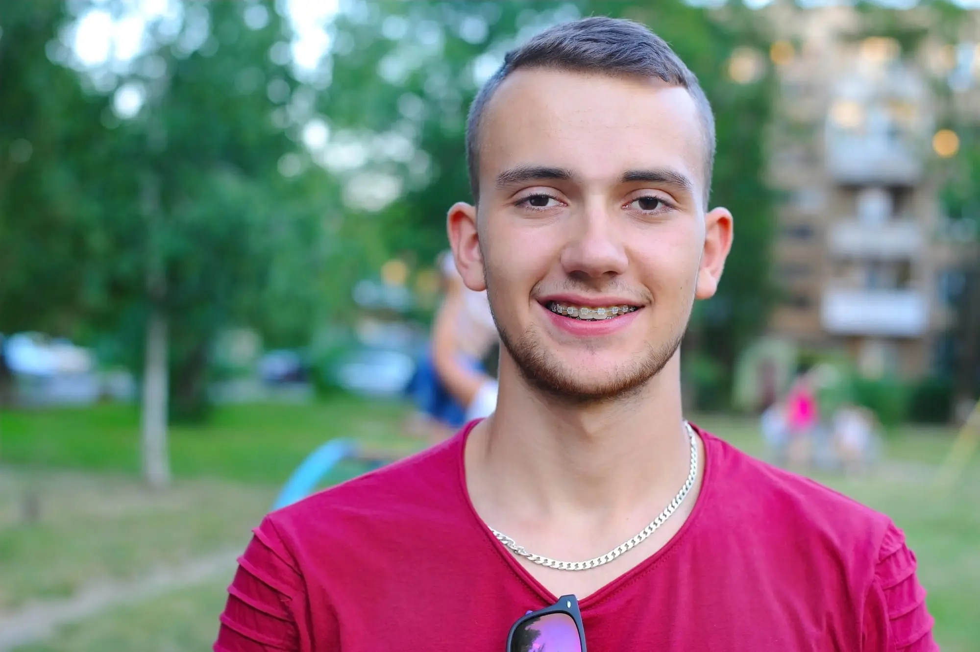 A young man with short hair and braces, red shirt, and chain necklace outside near trees and buildings, after visiting Forsyth Pediatric Dentistry and Orthodontics in Cumming, GA.