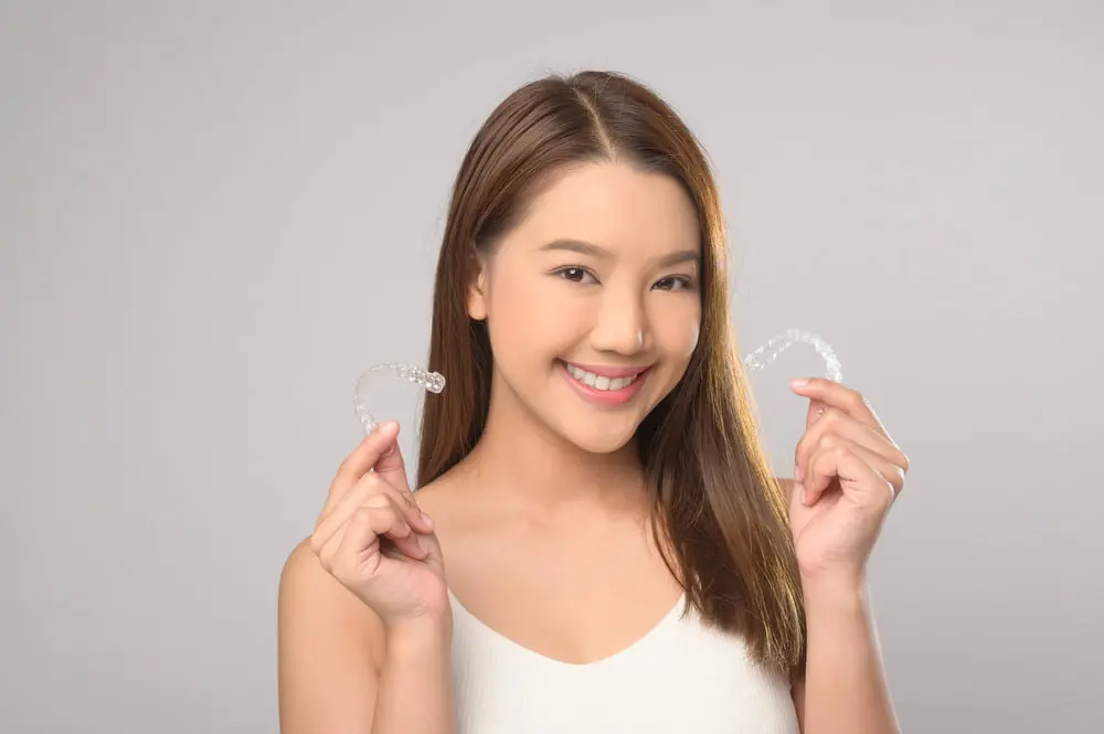 Smiling woman holds Invisalign aligners at Forsyth Pediatric Dentistry and Orthodontics in Cumming GA, with a plain background.
