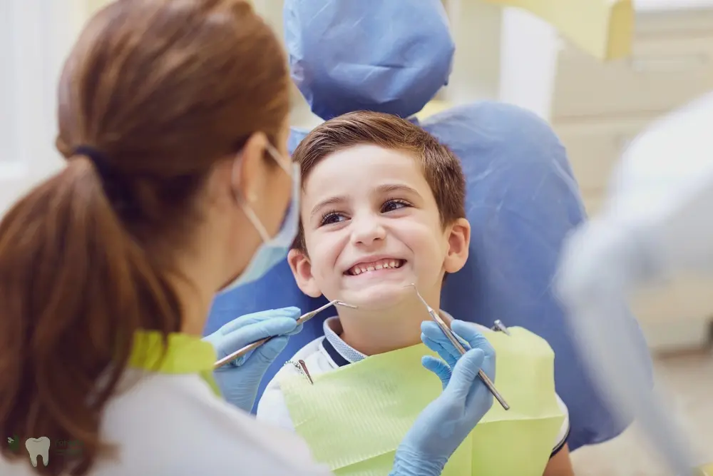 At Forsyth Pediatric Dentistry and Orthodontics in Cumming, GA, a masked dentist checks a smiling boy’s teeth with dental tools.