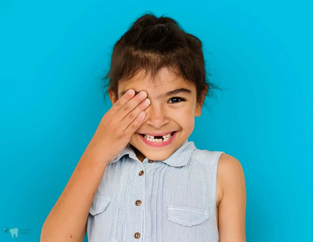 Smiling young child covers one eye, standing before a blue background for Forsyth Pediatric Dentistry and Orthodontics in Cumming, GA - Best Toothbrush for Kids by Age – Cumming Parent Guide