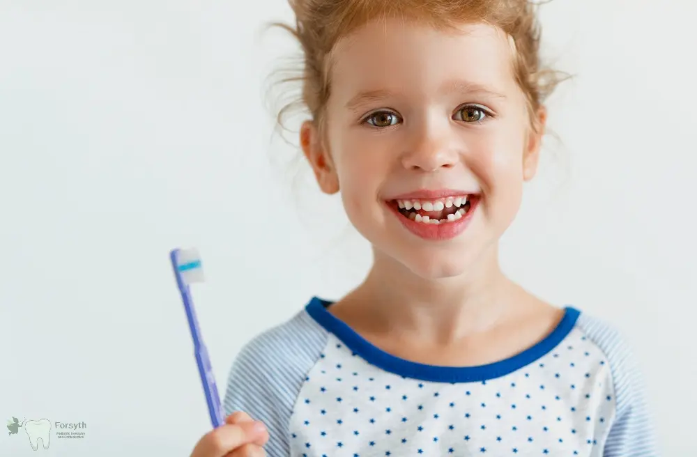 A smiling young girl with missing front teeth holds a toothbrush at Forsyth Pediatric Dentistry and Orthodontics in Cumming, GA - Fluoride Safety for Children in Cumming, GA