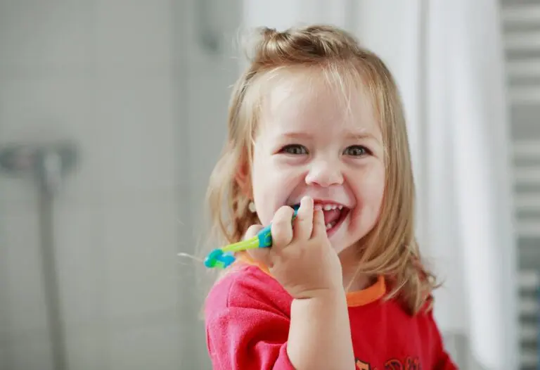 Smiling child with long hair in a red shirt brushes teeth in a bathroom at Forsyth Pediatric Dentistry and Orthodontics in Cumming, GA.