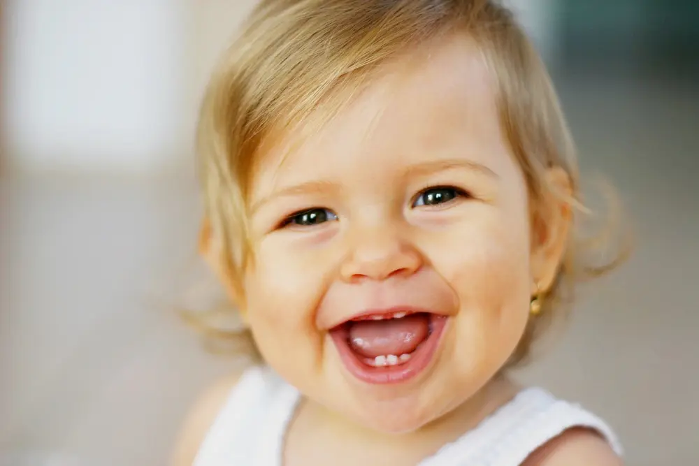 A smiling baby with light brown hair and a white outfit shows new teeth at Forsyth Pediatric Dentistry and Orthodontics in Cumming, GA.