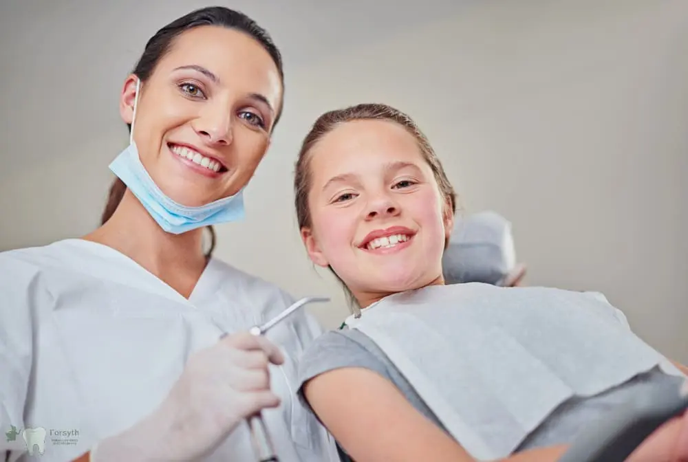 A dentist and young patient smile at the camera in a dental chair at Forsyth Pediatric Dentistry and Orthodontics in Cumming, GA - School Dental Health Programs in Cumming, GA 